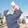 A person in a navy blue uniform and cap lifts a smiling infant dressed in a pink fleece jacket and white knit hat high into the air on what appears to be a maritime pier or dock. The joyful moment suggests a homecoming reunion, likely military in nature, given the uniformed personnel visible in the background. The scene takes place outdoors under a clear blue sky.