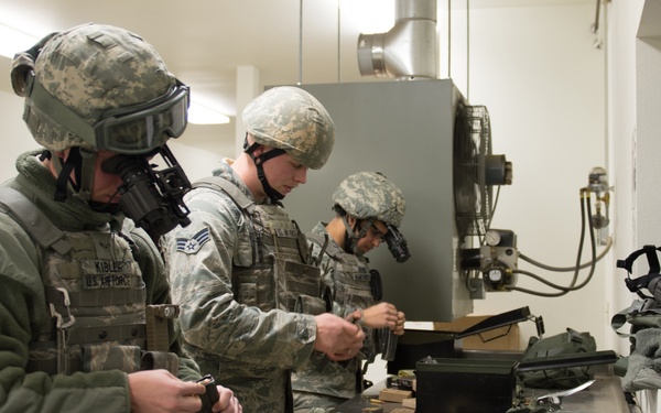 Three U.S. Air Force personnel in ACU/ABU camouflage uniforms and combat helmets are working at a metal counter inside what appears to be an armory or equipment staging area. The leftmost individual, identified by a name tape reading 'KIBLER' and 'U.S. AIR FORCE,' is wearing a gas mask. They appear to be loading magazines or conducting equipment checks with ammunition cans and gear spread across the counter.