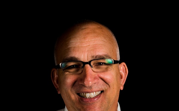 A professional portrait photograph of a middle-aged bald man smiling broadly against a solid black background. He is dressed in business-casual attire consisting of a white dress shirt and a bright blue tie. The image appears to be a formal headshot, likely used for professional or organizational purposes.