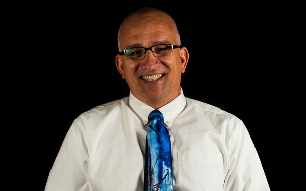 A bald man wearing glasses, a white dress shirt, and a decorative blue tie is seated at a dark surface with his hands clasped together. He is smiling broadly at the camera against a solid black background. The image appears to be a professional portrait photograph with studio lighting.