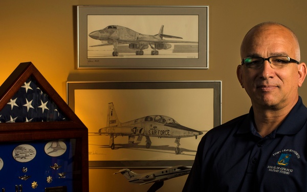 A middle-aged bald man wearing glasses and a navy blue polo shirt sits beside a wooden desk displaying military memorabilia. Behind him are framed pencil/charcoal drawings of military aircraft, including a U.S. Air Force jet. The setting appears to be a personal office or study with a warm amber lighting tone.