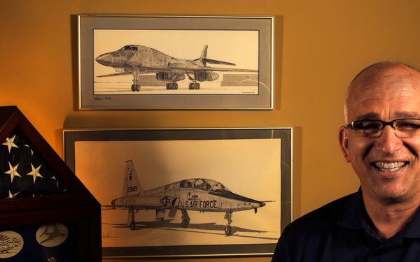 A smiling middle-aged man wearing a navy blue polo shirt with 'Fort Leavenworth School of Advanced Military Studies' insignia poses beside a military memorabilia display. The display includes a triangular flag case with an American flag and military medals/pins, aircraft artwork prints on the wall, and a small aircraft model on a stand. The setting appears to be a personal office or home study with military aviation memorabilia.