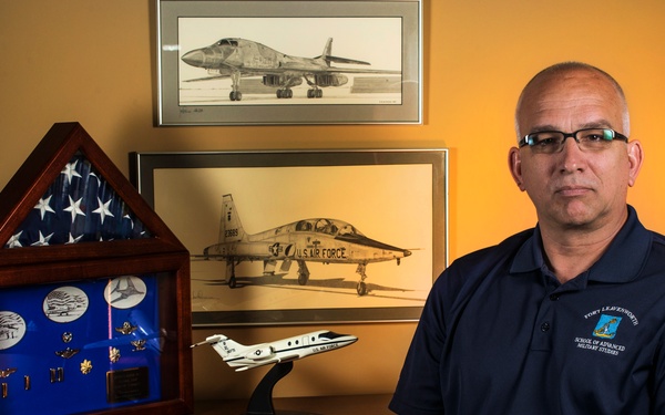 A middle-aged man wearing glasses and a navy blue 'Fort Leavenworth School of Advanced Military Studies' polo shirt sits in front of a wooden desk displaying military memorabilia. The display includes a triangular military shadow box with a folded American flag and medals, framed aircraft artwork, and a model aircraft. The setting suggests this is a military veteran or active service member's personal office or study.