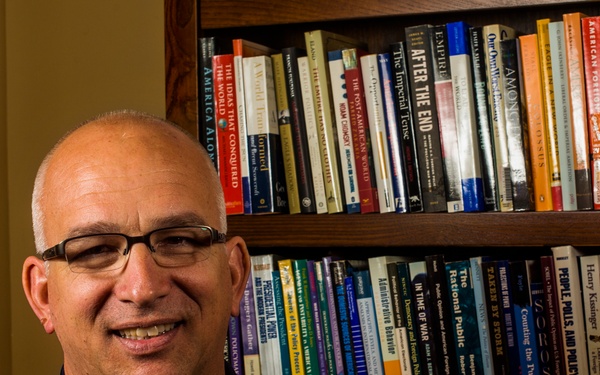 A middle-aged bald man wearing glasses and a navy blue polo shirt with a 'Fort Leavenworth School of Advanced' emblem is seated in front of a wooden bookshelf filled with numerous books. He is smiling and appears to be in an academic or professional office setting. The bookshelf behind him contains titles related to politics, foreign policy, military affairs, and international relations.