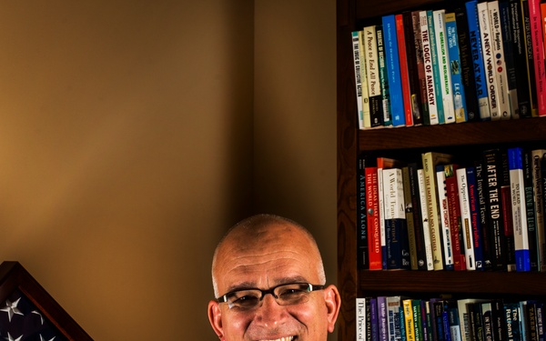 A bald man wearing glasses and a navy polo shirt bearing the 'Fort Leavenworth School of Advanced Military Studies' logo sits smiling in front of a large bookshelf filled with books on military and political topics. To his left is a triangular military shadow box display case containing a folded American flag, medals, and insignia on a blue background. The setting appears to be a personal office or study.