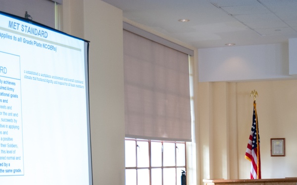 A U.S. Army soldier in ACU (Army Combat Uniform) is presenting a briefing in what appears to be a conference or training room. The presentation displayed on a projection screen covers the 'MET STANDARD' topic related to NCOERs (Non-Commissioned Officer Evaluation Reports). An American flag and a podium are visible in the background.