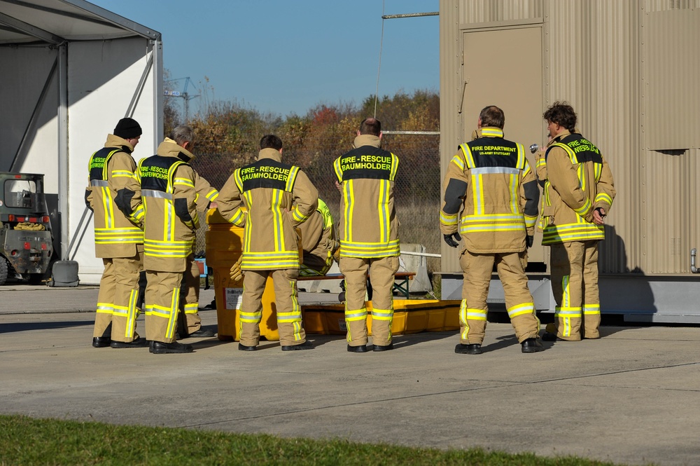 A group of firefighters and rescue personnel are gathered outdoors between two buildings, examining equipment on the ground. The personnel represent multiple units including Fire-Rescue Baumholder, Fire-Rescue Grafenwöhr, and Fire Department US Army Stuttgart. This appears to be a training exercise or joint operational briefing at a military installation.