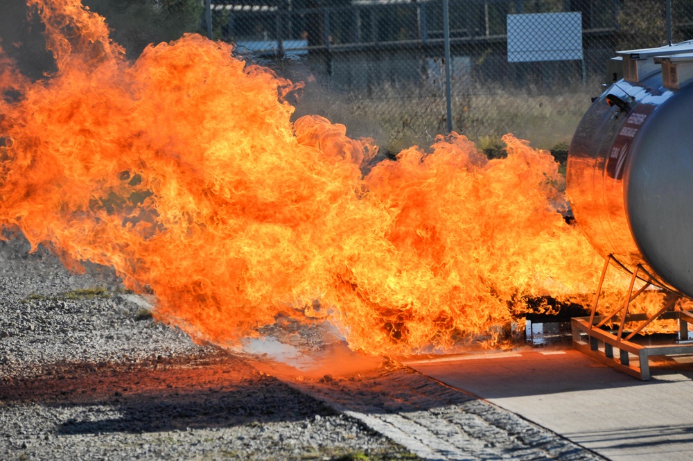 A large, intense fire is burning at ground level adjacent to a cylindrical industrial tank mounted on a metal support frame. The flames are predominantly orange and yellow, spreading horizontally to the left, suggesting a pressurized fuel or liquid release. The setting appears to be an industrial or training facility, visible by the chain-link fence and paved surface in the background.