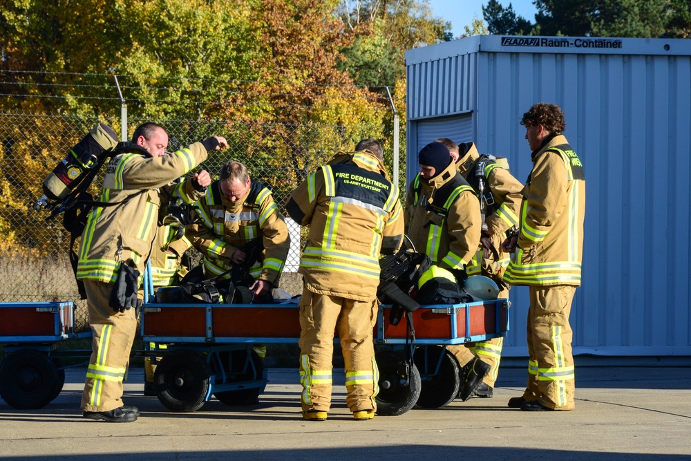 A group of firefighters in yellow turnout gear are gathered around a blue equipment cart, appearing to prepare or check gear. The text on one jacket reads 'FIRE DEPARTMENT US-ARMY STUTTGART', indicating this is a U.S. Army installation fire department in Stuttgart, Germany. The scene takes place outdoors near a chain-link fence and a blue storage container labeled 'FLADAFI Raum-Container', with autumn foliage visible in the background.