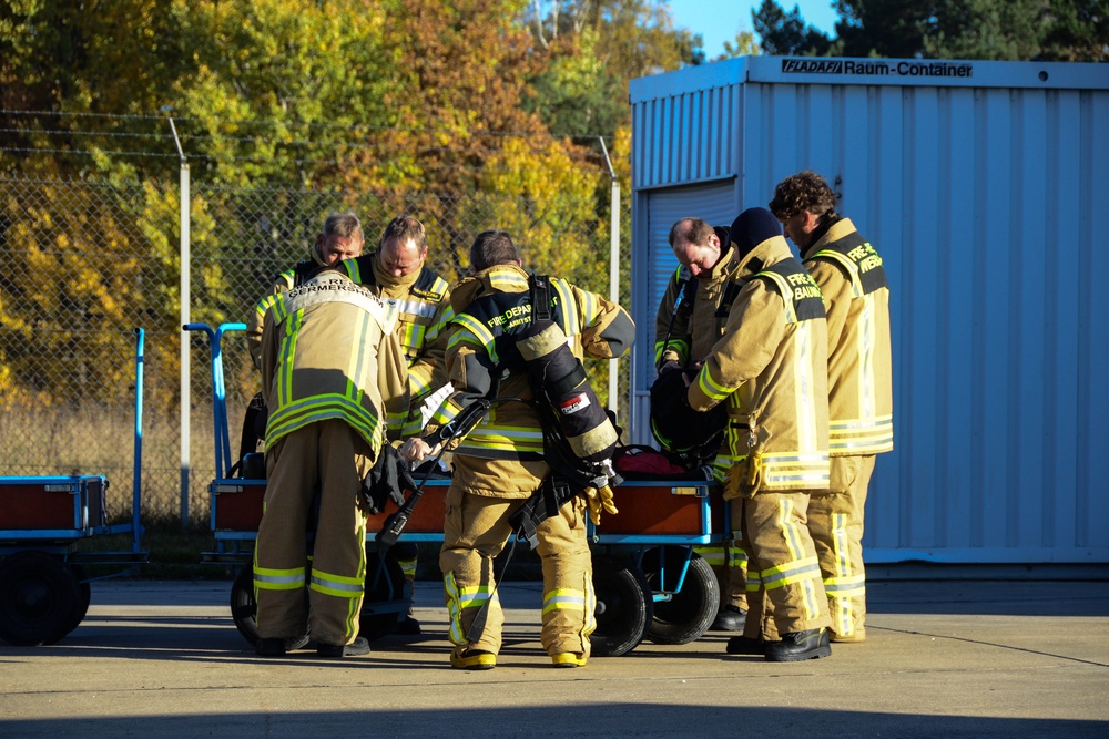 A group of approximately five firefighters in tan/yellow turnout gear are gathered around a wheeled stretcher or cart in an outdoor paved area. They appear to be conducting a training exercise or preparing equipment, with one firefighter wearing a self-contained breathing apparatus (SCBA). The setting includes a chain-link fence, autumn trees, and a blue modular container labeled 'FLADAFI Raum-Container' in the background.