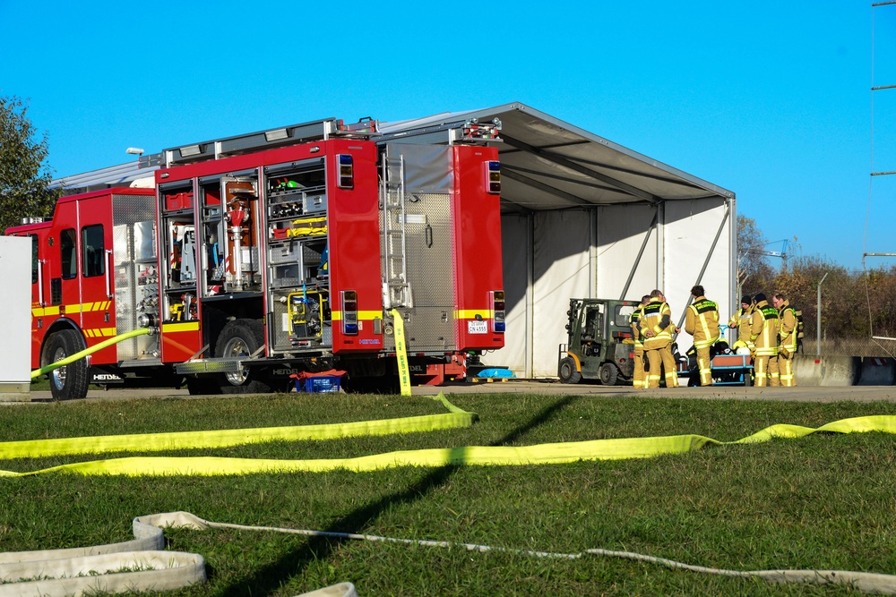 A red fire truck with its equipment compartments open is parked near a large temporary white structure or hangar. Several firefighters in yellow protective gear are gathered near the entrance of the structure alongside a forklift. Yellow fire hoses are deployed across the foreground grass, suggesting an active training exercise or emergency response operation.