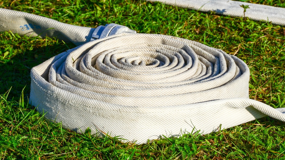 A white canvas fire hose is tightly coiled in a flat spiral pattern and resting on green grass. The hose appears to be a flat-lay type, consistent with firefighting equipment, and shows signs of use with visible dirt and wear. A blue identification tag or strap is visible near the top of the coil.