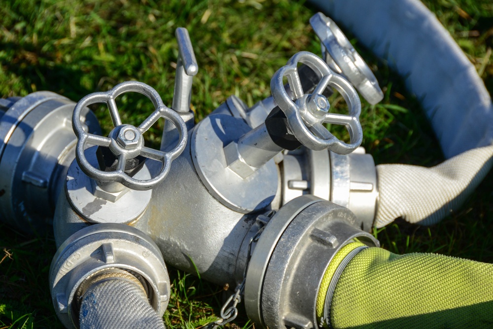 The image shows a close-up of a firefighting water distribution manifold (verteiler) with multiple valve outlets resting on green grass. Two handwheel gate valves are clearly visible on top of the silver aluminum assembly, with multiple hose couplings attached. Several fire hoses in different colors (gray, blue, and yellow-green) are connected to the manifold.