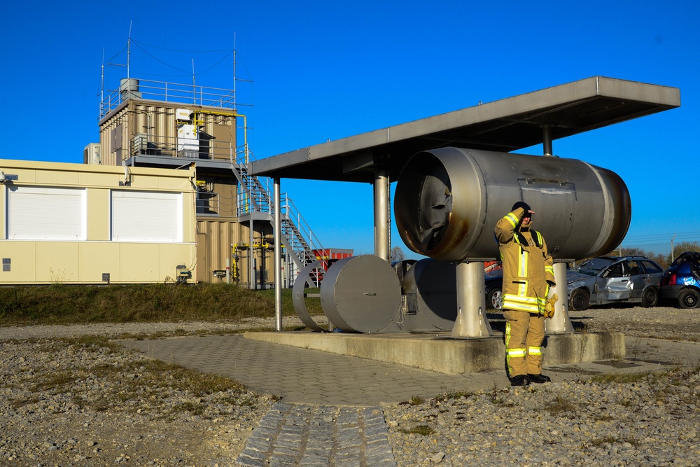 The image shows a firefighter in full protective gear standing beside large industrial cylindrical tanks or vessels at what appears to be a fire training facility or industrial complex. The facility includes a multi-story training tower with stairs and platforms in the background, along with various industrial equipment. The scene is set on a clear, sunny day with bright blue skies.
