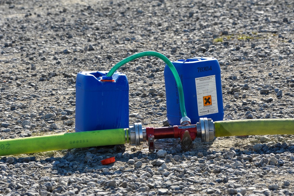 The image shows two blue plastic jerry cans connected via a green flexible hose to a red metal valve/manifold junction, which is also connected to a larger green fire hose lying on a gravel surface. The right jerry can bears a label reading 'ÜBUNGSSCHAUMMITTEL U / TRAININGFOAM U' (Training Foam U) with a hazard symbol (harmful/irritant - orange X symbol), and the number '70304' is visible. This appears to be a firefighting training exercise setup using foam concentrate.