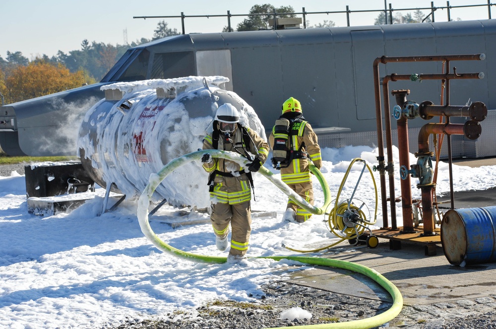 Two firefighters in full protective gear are conducting what appears to be a firefighting training exercise, applying foam suppressant to a tank or vessel. The ground and target object are heavily covered in white firefighting foam. The scene takes place at what appears to be a dedicated fire training facility with industrial pipe infrastructure visible.