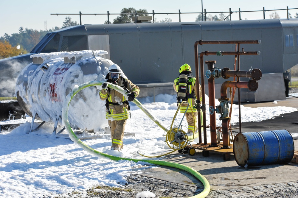 Two firefighters in full protective gear and breathing apparatus are applying foam suppressant to a tank or vessel at what appears to be a fire training facility. The tank is heavily covered in white firefighting foam, and the scene includes industrial piping infrastructure and a blue drum nearby. This appears to be a live fire training exercise conducted at a dedicated training ground.