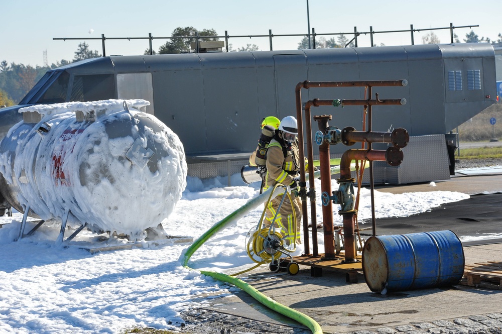 Two firefighters in full protective gear and breathing apparatus are operating at what appears to be a fire training facility or industrial site. They are working with a green hose connected to a pipe manifold system, while a large tank or vessel is covered in white firefighting foam. The scene suggests an active firefighting exercise or emergency response involving foam suppression.
