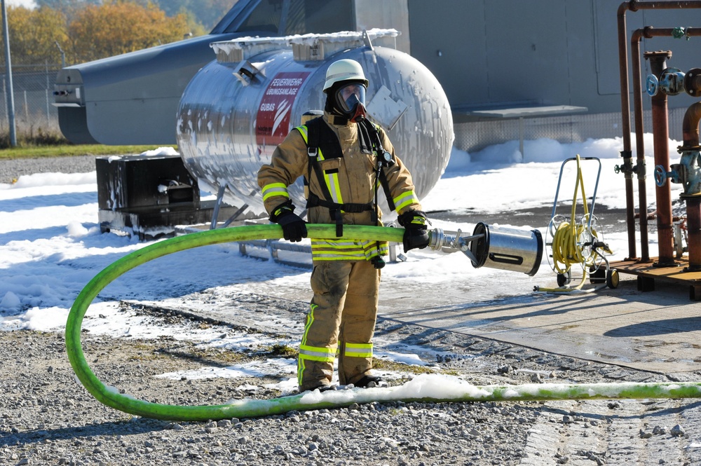 A firefighter in full protective gear, including a respirator mask and white helmet, is holding a green fire hose with a metal nozzle at what appears to be a fire training facility. The ground is covered with fire suppression foam, and a large silver tank with a sign reading 'Feuerwehr Übungsanlage GUBAS' (Fire Department Training Facility GUBAS) is visible in the background. The scene is consistent with a controlled firefighting exercise or drill.