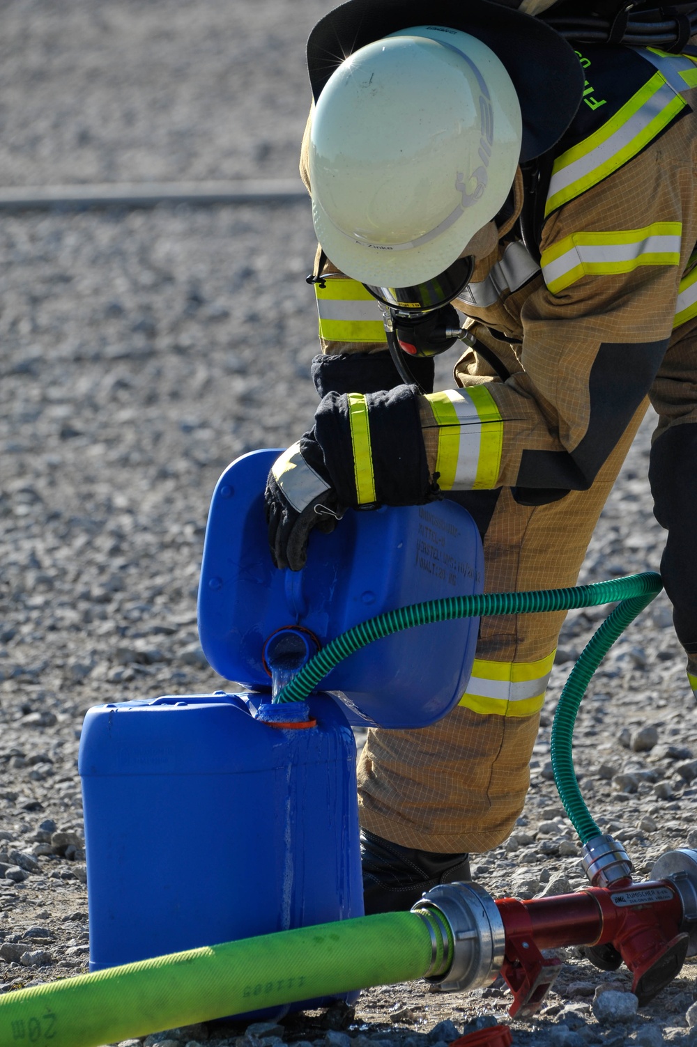 A firefighter in full turnout gear, including a white helmet and breathing apparatus, is bent over handling blue plastic containers connected to green hoses. The scene appears to be a training exercise or hazmat operation taking place on a gravel surface. The firefighter is manipulating what appears to be a liquid transfer or foam injection setup.