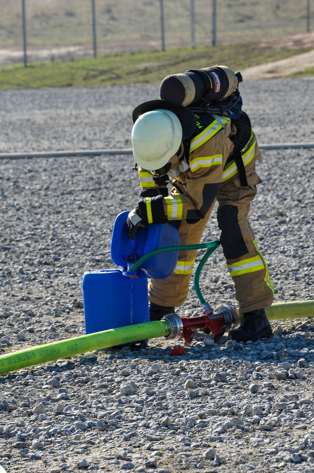 A firefighter in full turnout gear, including SCBA (self-contained breathing apparatus), is bent over working with hose connections and a blue liquid container on a gravel surface. The scene appears to be an outdoor training exercise or operational scenario. Green fire hoses are connected to a red manifold/wye fitting on the ground.