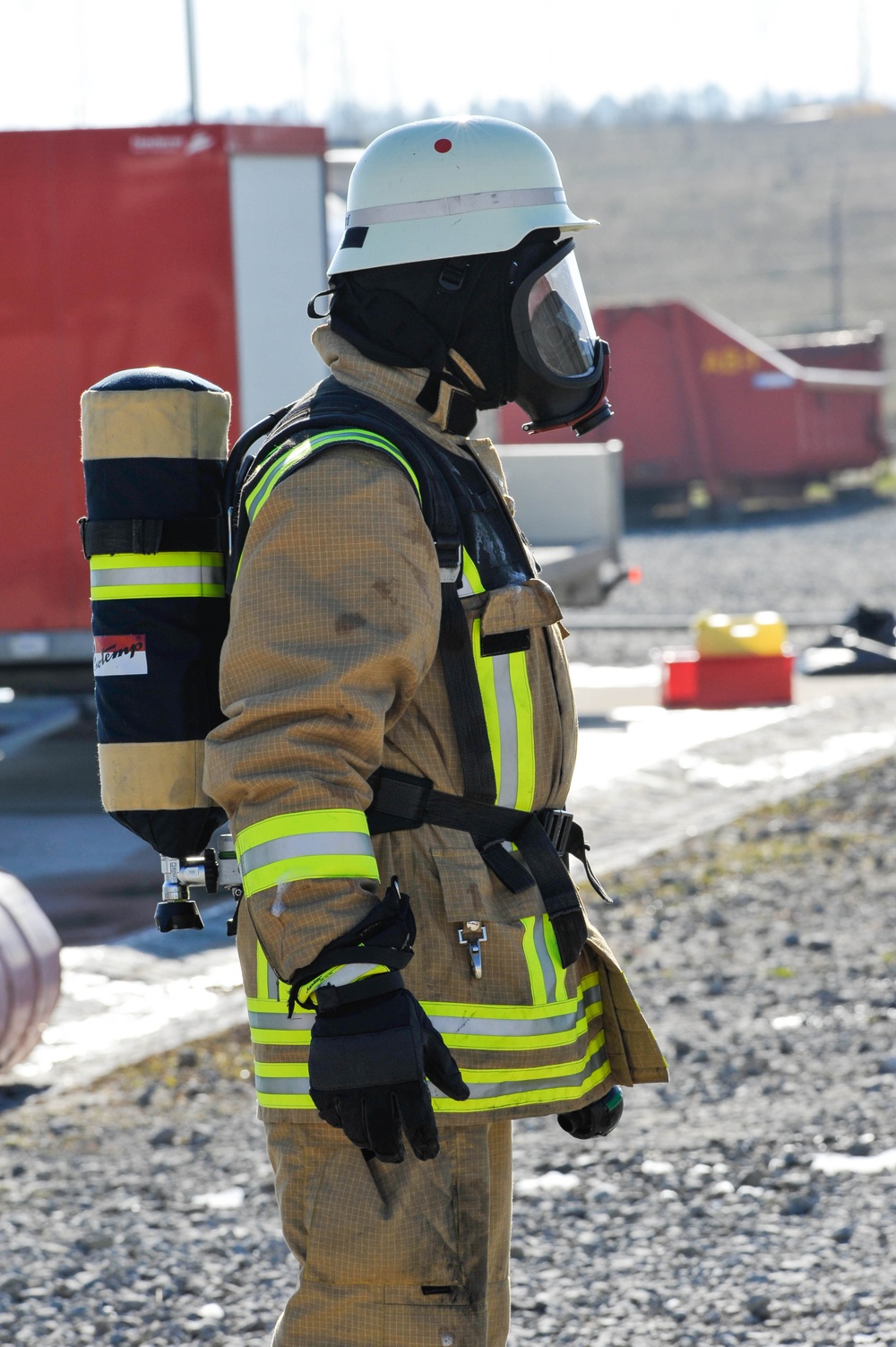 A fully equipped firefighter stands on a gravel surface at what appears to be a training facility or emergency response site. The individual is wearing full personal protective equipment including a turnout suit, SCBA (Self-Contained Breathing Apparatus), full-face respirator mask, white helmet, and black gloves. The background shows red containers, equipment, and an open outdoor industrial area.