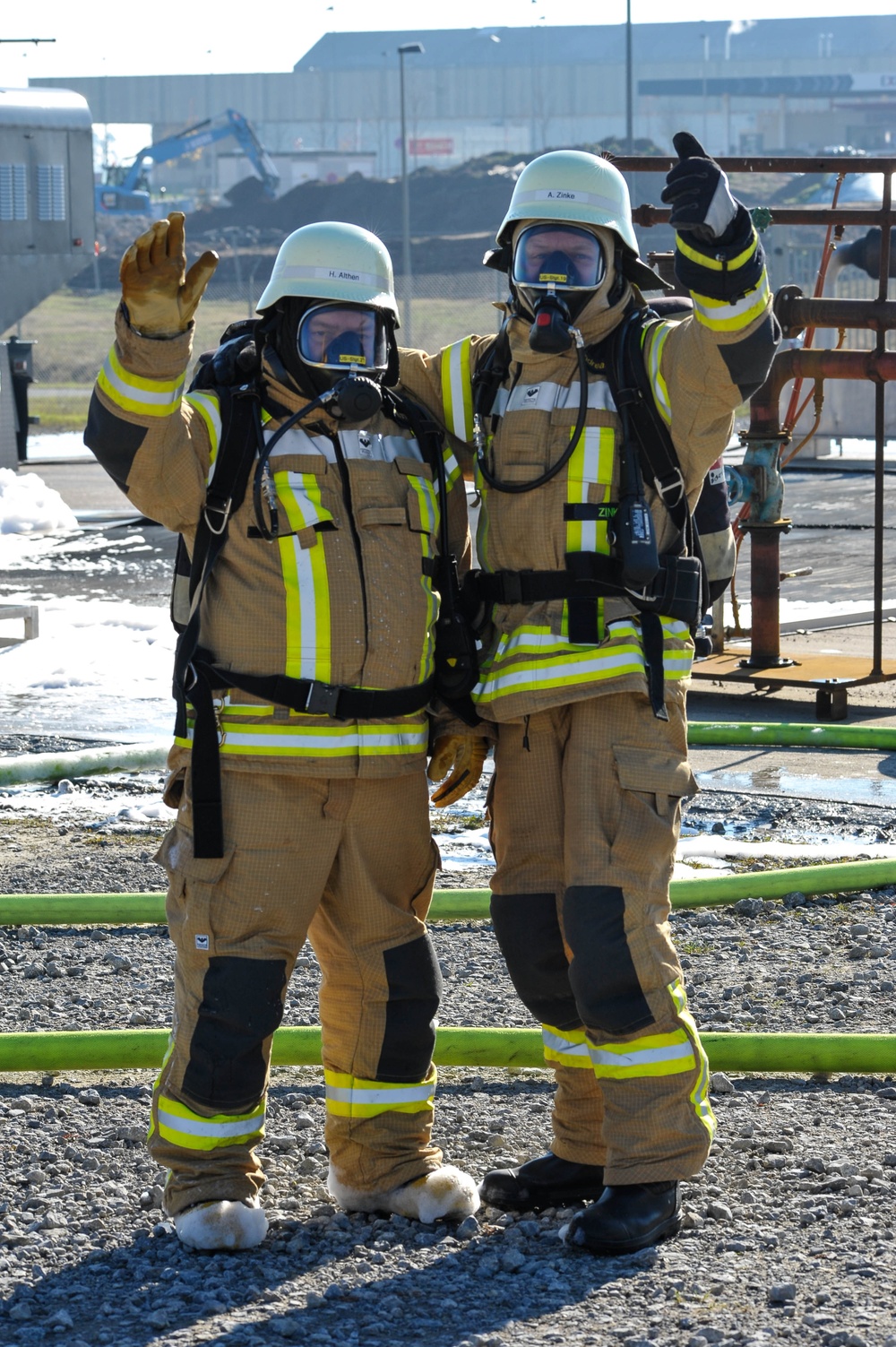 Two firefighters in full personal protective equipment, including breathing apparatus and full-face respirator masks, pose together at what appears to be an industrial training or incident site. Both are wearing tan/gold turnout gear with high-visibility yellow-green reflective striping and white helmets with name labels visible ('H. Althen' and 'A. Zinke'). They are giving thumbs-up and wave gestures, suggesting successful completion of a drill or operation.