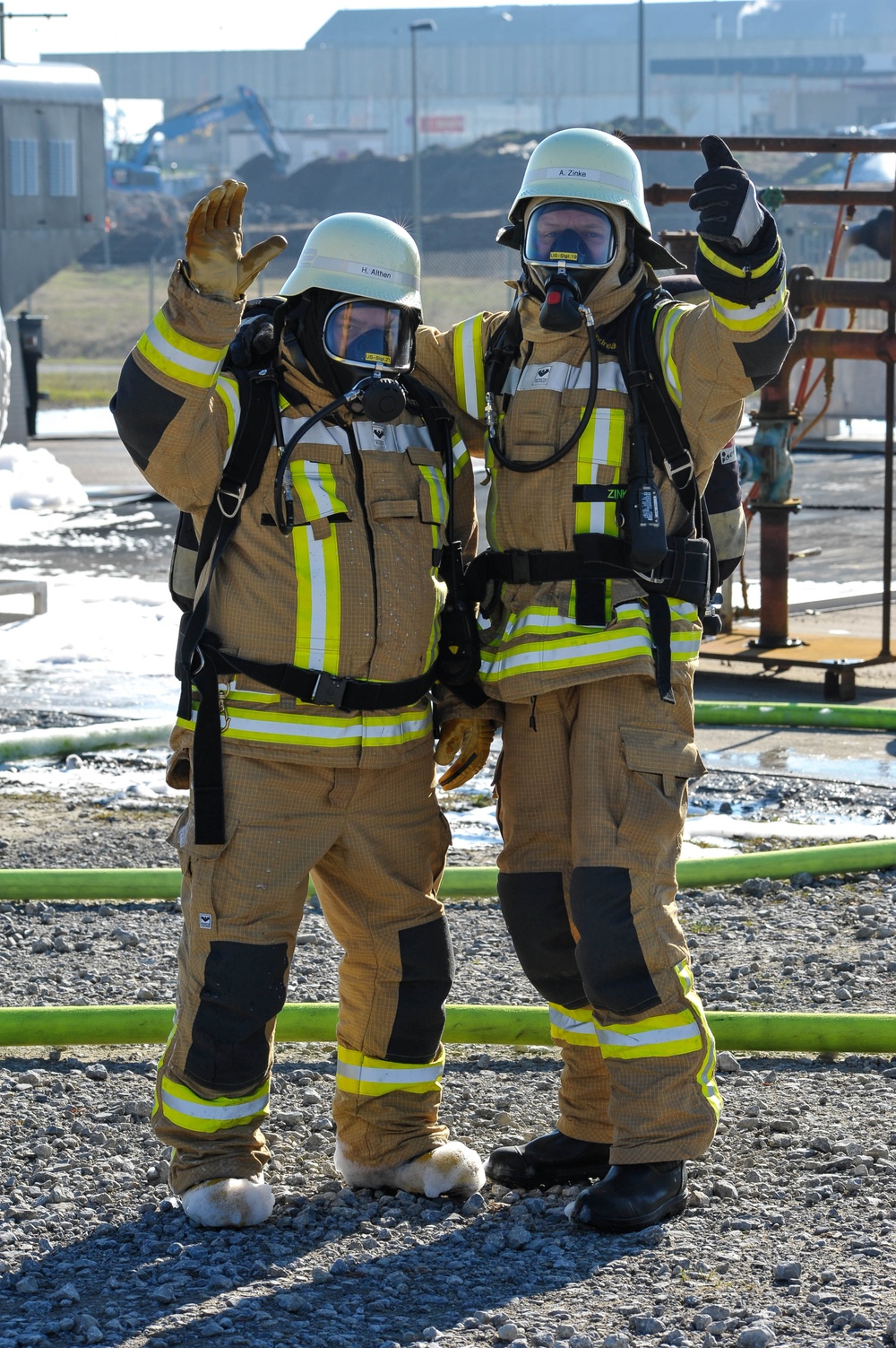 Two firefighters in full personal protective equipment pose together at what appears to be an industrial training or incident site. Both are wearing tan/gold turnout gear with high-visibility yellow and white reflective striping, white helmets, full-face respirator masks, and self-contained breathing apparatus (SCBA). They are giving hand signals — one raising an open hand and the other giving a thumbs-up — suggesting a successful completion of a drill or operation.