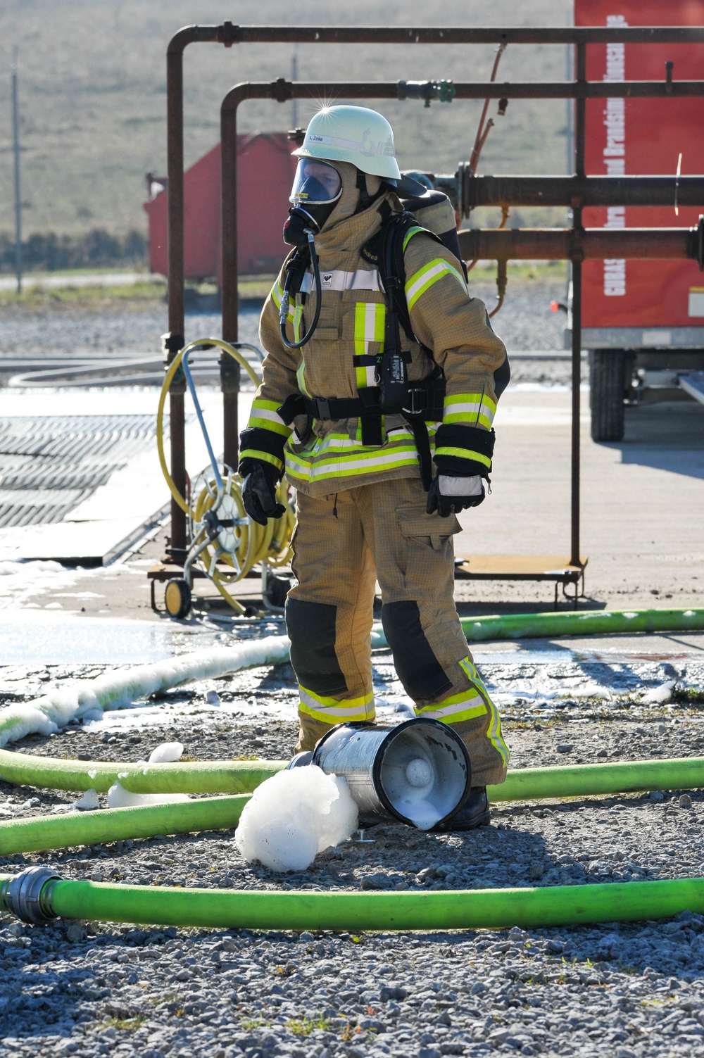 A firefighter in full personal protective equipment stands at what appears to be a training exercise or incident site. The firefighter is wearing a tan/gold turnout gear with high-visibility yellow reflective stripes, a white helmet, and a full-face SCBA (Self-Contained Breathing Apparatus) mask. Green fire hoses are laid across a gravel surface, and a metal container with white foam/suppressant agent is visible at the firefighter's feet.