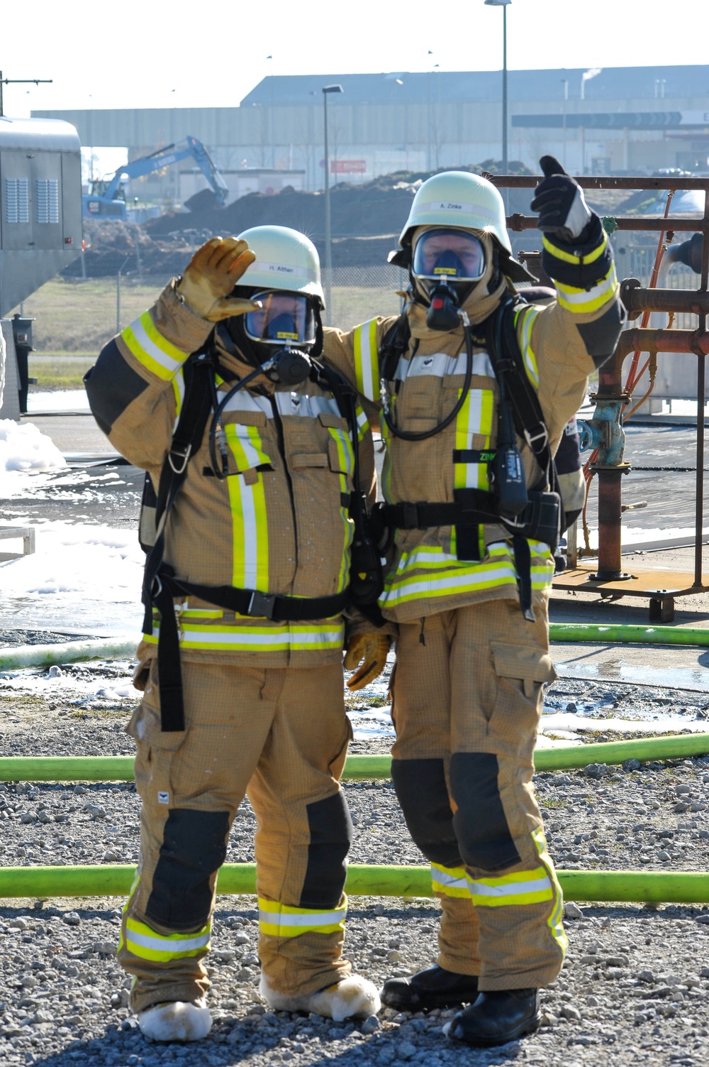 Two firefighters in full personal protective equipment are posing together at what appears to be an industrial training or incident site. Both are wearing breathing apparatus, helmets with name labels, and high-visibility turnout gear with yellow/green reflective striping. They are giving thumbs-up and salute gestures, suggesting a successful completion of a task or exercise.