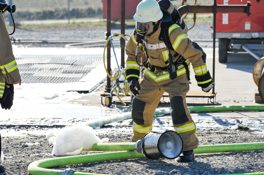 A firefighter in full personal protective equipment (PPE), including a white helmet, SCBA (self-contained breathing apparatus), and tan turnout gear with high-visibility reflective striping, is working at what appears to be a fire training or incident scene. The ground is covered with firefighting foam and green hoses are laid out across a gravel surface. A second firefighter is partially visible on the left edge of the frame.