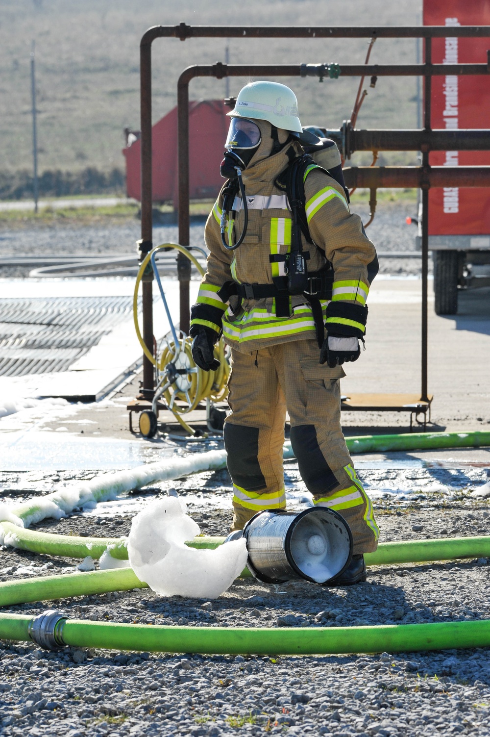A firefighter in full personal protective equipment stands at what appears to be a training facility or active incident scene. The firefighter is wearing a tan/gold turnout gear with high-visibility yellow-green reflective strips, a white helmet, and a full-face SCBA (Self-Contained Breathing Apparatus) mask. Green fire hoses are laid across a gravel surface, and firefighting foam is visible near a toppled metal container.