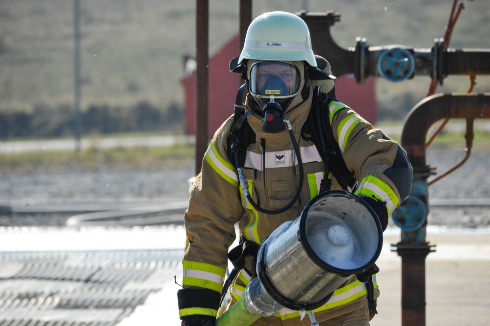 A firefighter in full personal protective equipment is captured in an outdoor industrial setting, likely during a training exercise or active response. The individual is wearing a tan/gold turnout gear with high-visibility yellow reflective stripes, a white helmet labeled 'A. Zinke', and a full-face SCBA (Self-Contained Breathing Apparatus) mask. They are carrying what appears to be a foam or dry chemical fire suppression nozzle/applicator.