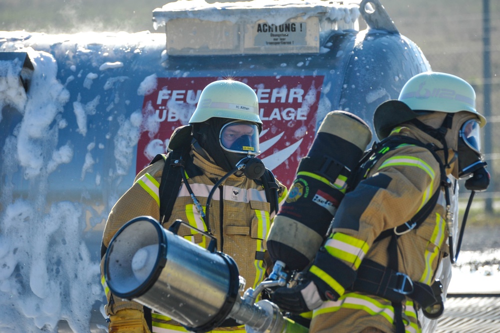 Two German firefighters in full protective gear including breathing apparatus and helmets are operating at what appears to be a fire training facility (Feuerwehr Übungsanlage). They are handling firefighting equipment, likely a foam or CO2 extinguisher nozzle, with foam/suppression agent visible covering a large tank or vessel behind them. The scene depicts an active firefighting exercise or real incident response.