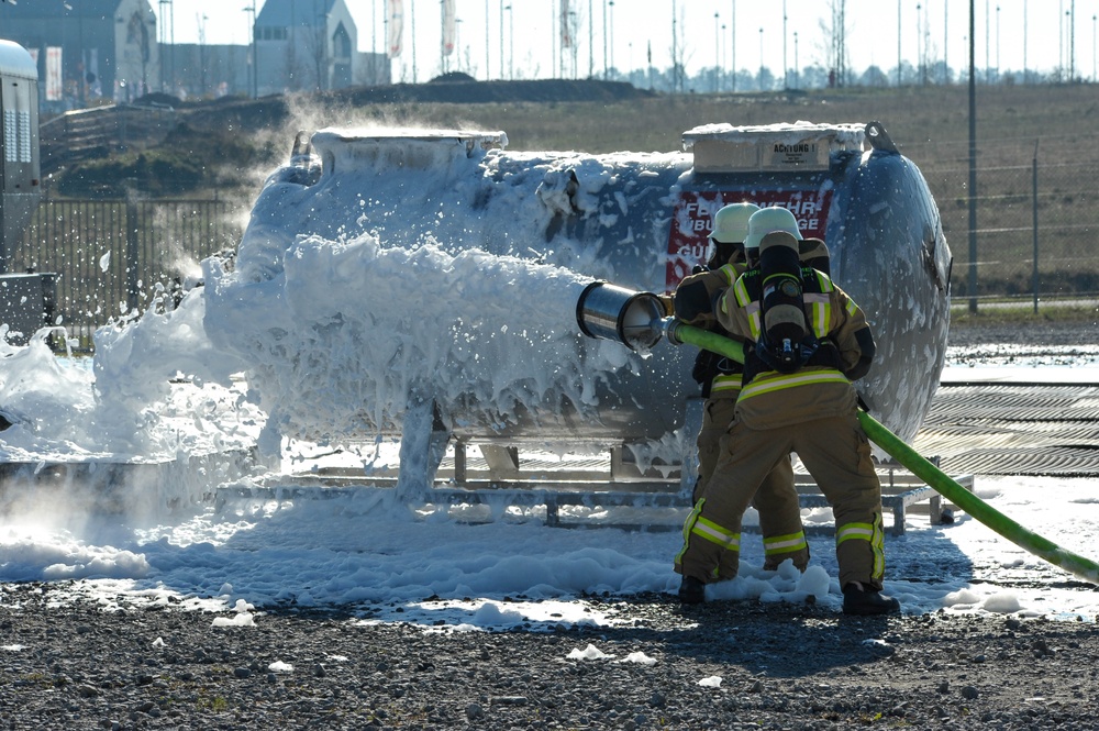 Two firefighters in full protective gear and self-contained breathing apparatus (SCBA) are applying foam suppression agent onto a large industrial tank using a hose. The scene appears to be a firefighting training exercise, as indicated by the sign in the background containing the word 'FEUERWEHR' (German for 'fire department'). Large amounts of white foam are covering the tank and spreading across the gravel ground.
