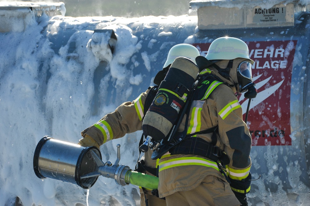 Two firefighters in full protective gear, including SCBA (self-contained breathing apparatus) tanks and reflective turnout suits, are operating a foam suppression nozzle. The scene shows a large amount of firefighting foam covering a structure or container in the background. A German-language warning sign reading 'ACHTUNG' and partial text 'WEHR ANLAGE' is visible on the right side.