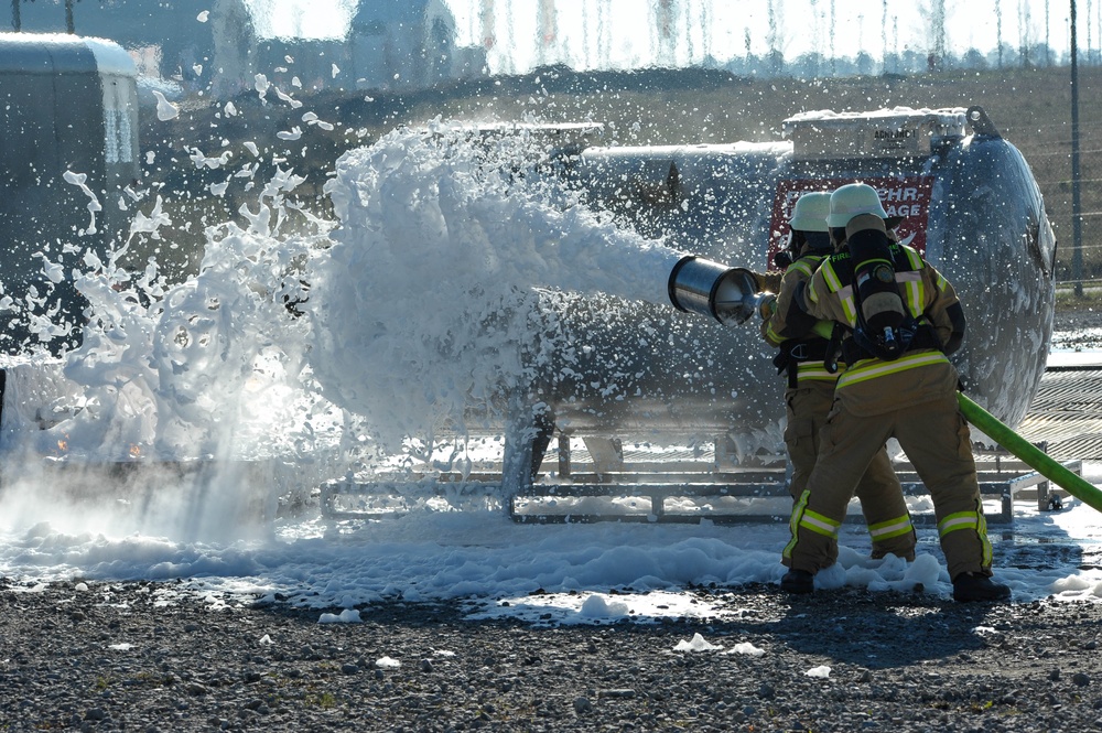 Two firefighters in full protective gear and breathing apparatus are directing a high-pressure foam hose at what appears to be a tank or industrial vessel. The foam is dispersing dramatically upon impact, creating a large white spray cloud. This appears to be either a live fire suppression operation or a training exercise at an industrial facility.