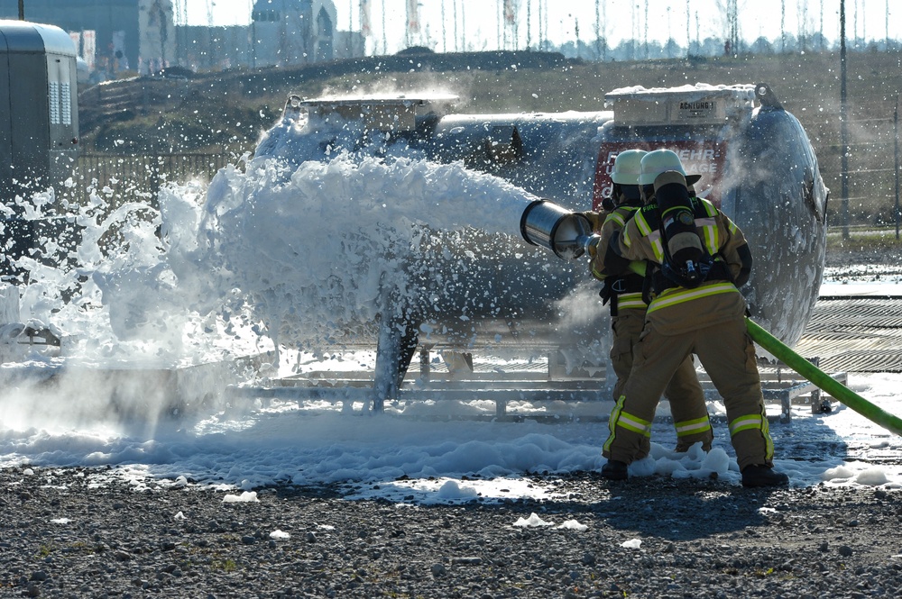 Two firefighters in full protective gear and breathing apparatus are directing a foam suppression hose at what appears to be a pressurized tank or vessel. White firefighting foam is being discharged in large quantities, covering the ground and the target structure. This appears to be either an active incident response or a training exercise involving a hazardous material or fuel tank.