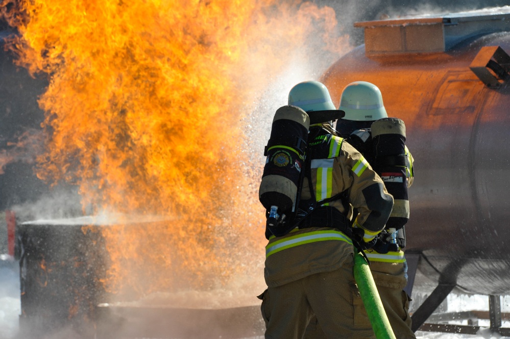 Two firefighters in full protective gear are actively combating a large fire using a green hose. The fire appears to be emanating from an industrial area, with a large orange tank or vessel visible in the background. Water and steam are visible as the firefighters direct their hose toward the base of the flames.