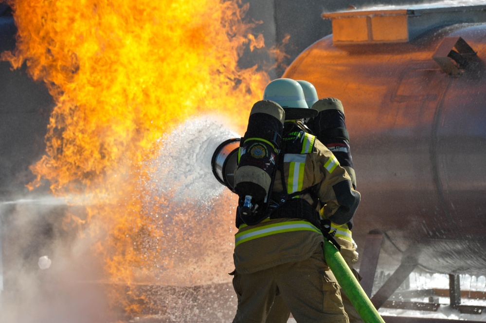 Two firefighters in full protective gear are actively combating a large open flame using a fire hose. The fire appears to be emanating from or near an industrial tank or vessel visible in the background. The image captures an intense firefighting operation with water/foam spray visibly engaging the flames.