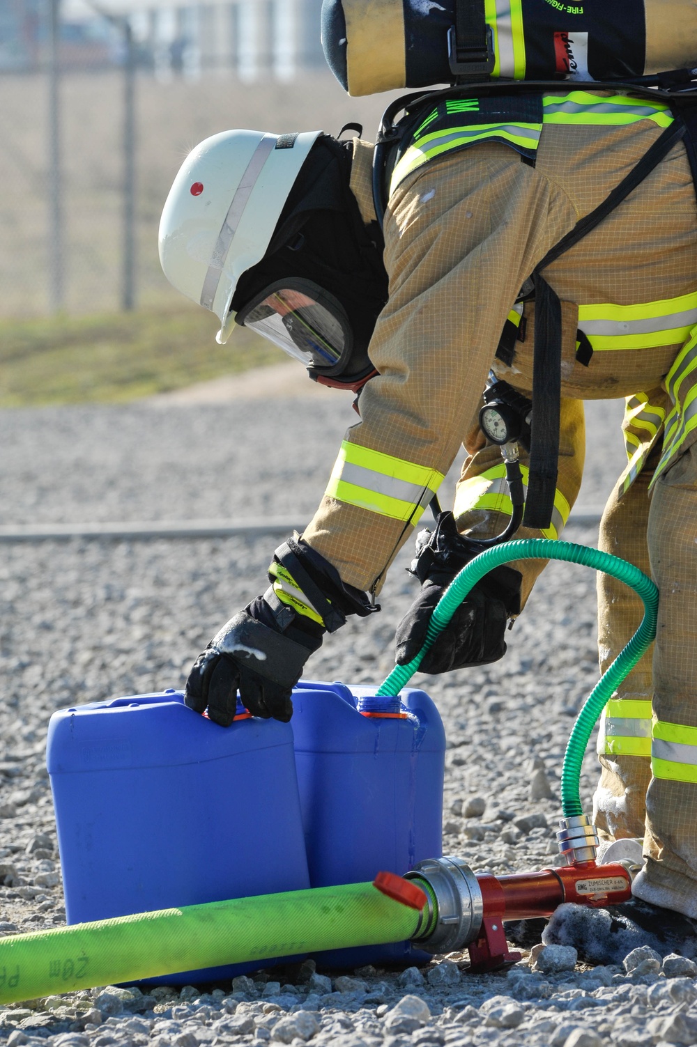 A firefighter in full personal protective equipment, including a white helmet, SCBA (self-contained breathing apparatus), and turnout gear with high-visibility yellow-green reflective stripes, is bending down to handle equipment on a gravel surface. The firefighter appears to be managing a blue container and green hose connected to a red-handled nozzle assembly. This appears to be a training exercise or operational deployment at an outdoor site near a fenced perimeter.