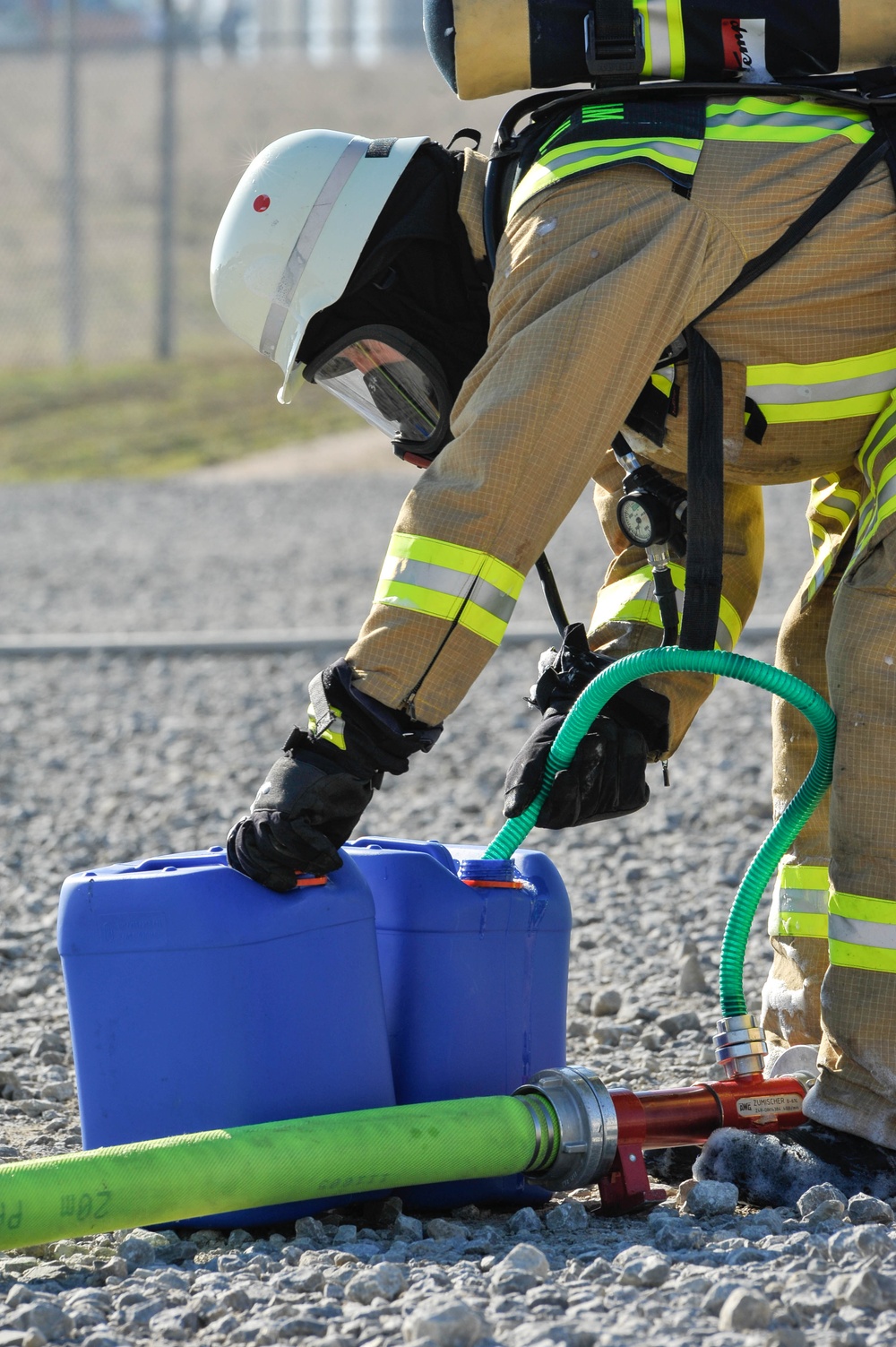 A firefighter in full personal protective equipment, including a breathing apparatus and white helmet, is bent over handling a blue plastic container on a gravel surface. Green hoses are connected to a red manifold/valve assembly near the container. This appears to be a hazmat or firefighting training exercise or operational scenario.