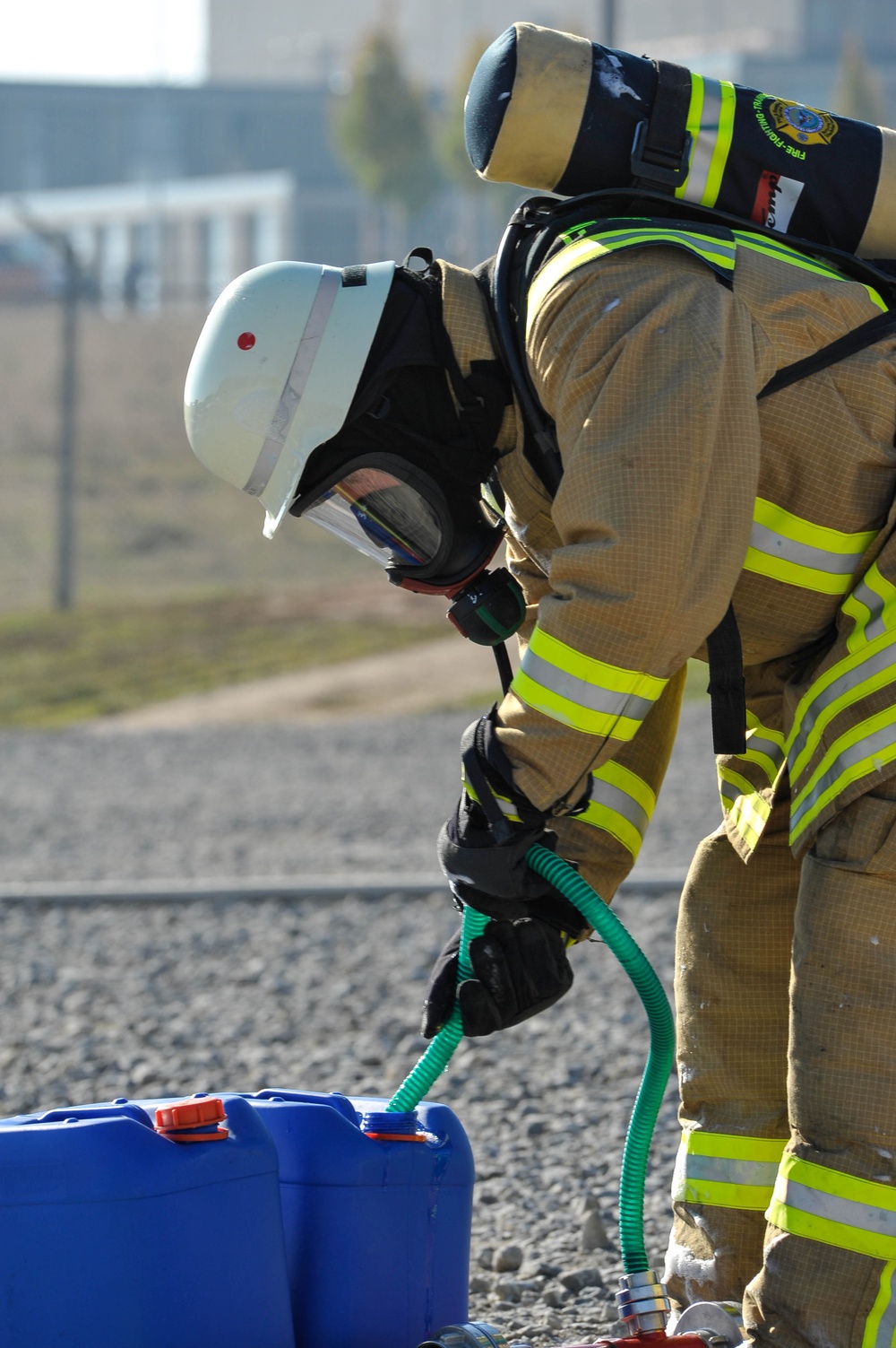 A firefighter in full personal protective equipment is bent over a blue plastic container, handling a green corrugated hose. The individual is wearing a tan/gold turnout gear with high-visibility yellow-green reflective stripes, a white helmet, full-face SCBA mask, and a self-contained breathing apparatus (SCBA) tank on their back. The setting appears to be an outdoor training or operational area with gravel ground and a fenced facility in the background.