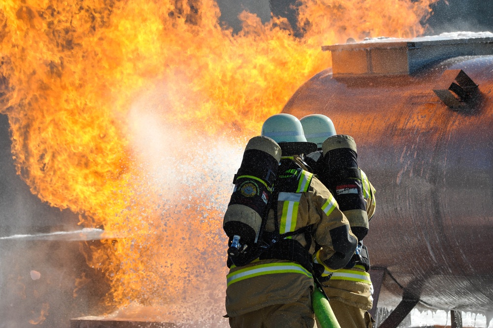 Two firefighters in full protective gear are actively combating a large fire near an industrial tank or vessel. They are working in tandem, directing a water or foam stream toward intense orange flames. The scene appears to be an industrial fire emergency or possibly a training exercise involving a fuel or chemical storage tank.