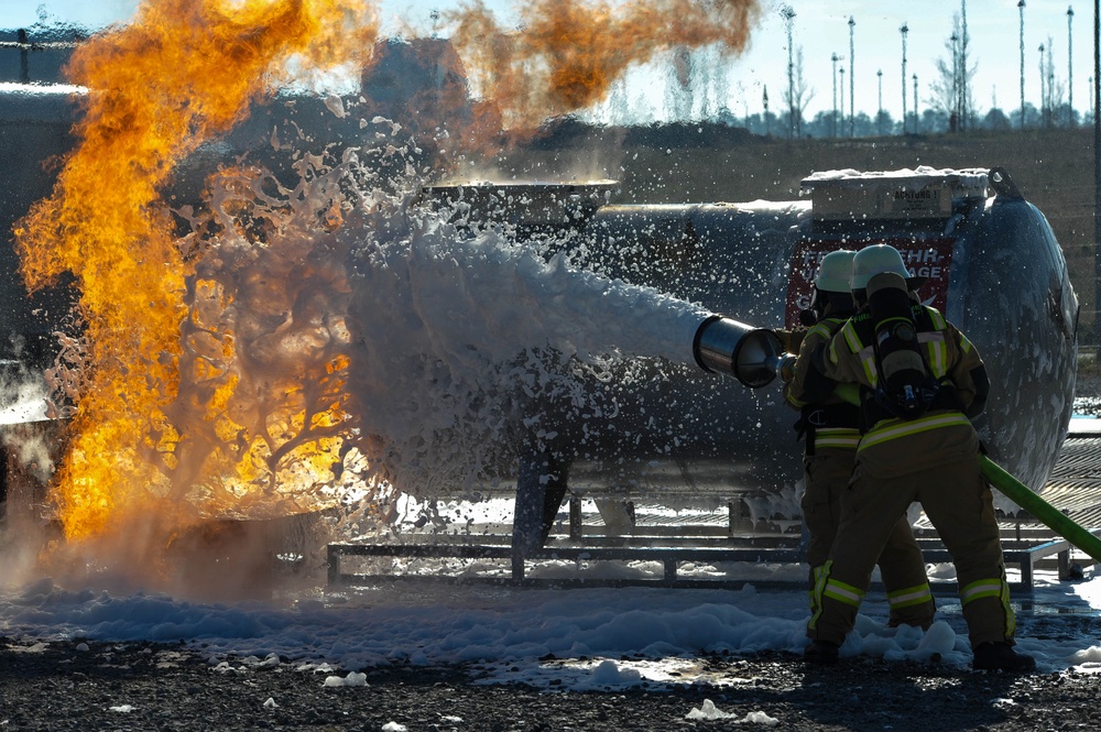 Two firefighters in full protective gear are actively combating a large fuel or chemical fire using a foam suppression hose. The foam agent is visibly colliding with the intense orange flames, creating a dramatic splash effect. The setting appears to be an industrial training or emergency response facility.