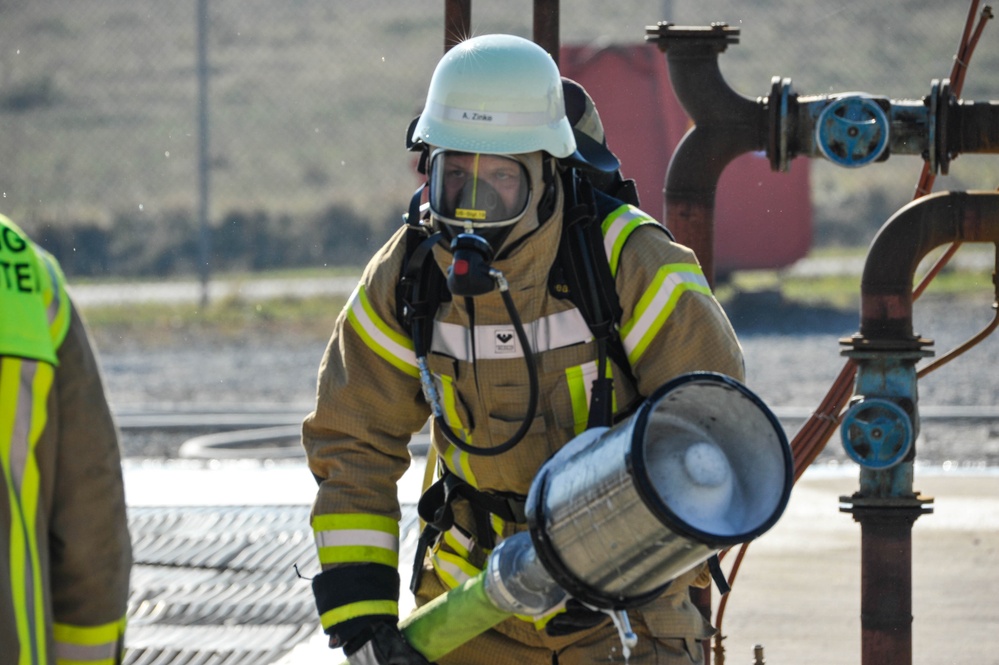 A firefighter in full personal protective equipment, including a tan/gold turnout gear with high-visibility yellow reflective stripes, a white helmet labeled 'A. Zeke', and a full-face SCBA (Self-Contained Breathing Apparatus) mask, is carrying a foam or dry chemical fire extinguisher nozzle. The setting appears to be an industrial facility or fire training ground, with visible pipe infrastructure and valves in the background. A second person in a high-visibility green vest is partially visible on the left edge of the frame.