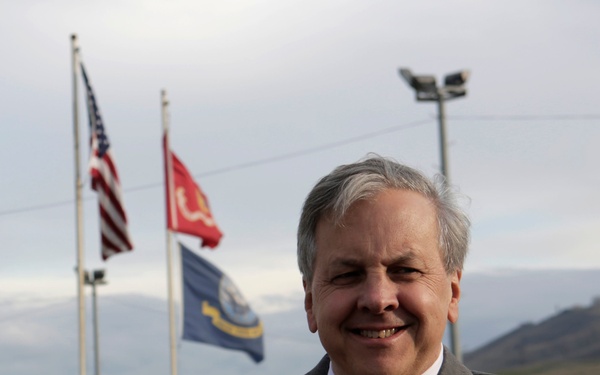 A middle-aged man in a dark suit and patterned tie stands with arms crossed, smiling, at what appears to be a military installation. Three flags are visible in the background, including the American flag and what appears to be a Marine Corps flag. A person in military camouflage uniform is visible in the background.