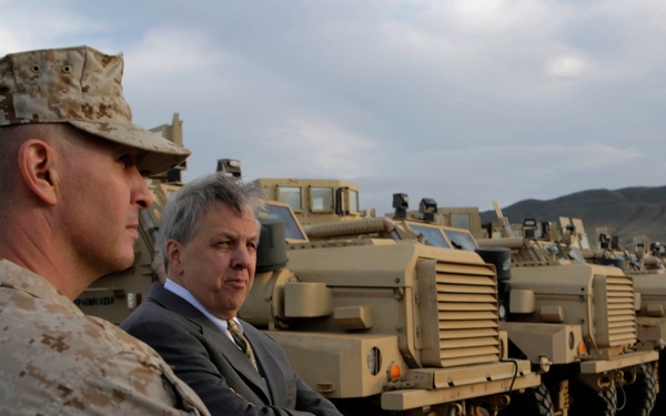 Two individuals — a military officer in desert camouflage uniform with a patrol cap and a civilian in a dark suit — stand before a row of tan/sand-colored military armored vehicles lined up in a desert or arid terrain setting. The vehicles appear to be MRAP-type (Mine-Resistant Ambush Protected) trucks arranged in a formal display or inspection line. Mountains and an overcast sky are visible in the background.