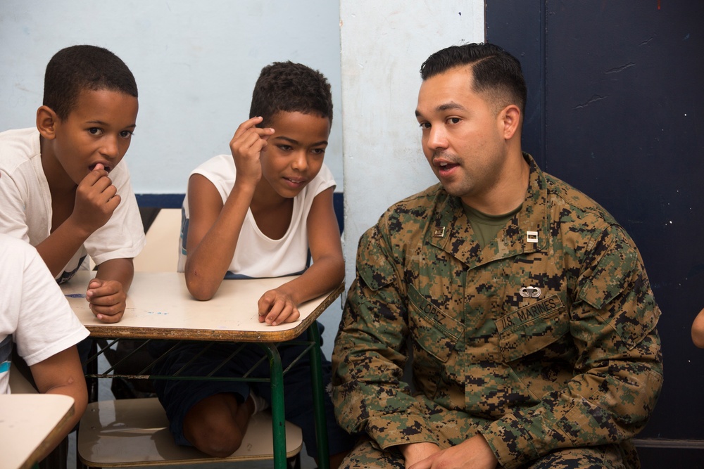 A U.S. Marine in woodland MARPAT camouflage uniform is seated and speaking with two young boys in what appears to be a classroom setting. The Marine, whose name tape reads 'LOPEZ' and who wears U.S. Marines insignia, appears to be engaged in a community outreach or educational visit. The children are attentively listening and interacting with the Marine at a school desk.