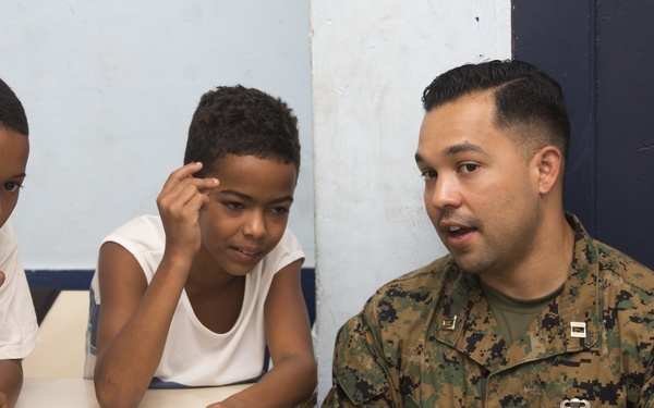 A U.S. Marine in woodland MARPAT camouflage uniform is seated and speaking with two young boys in what appears to be a classroom setting. The Marine, whose name tape reads 'LOPEZ' and who wears U.S. Marines insignia, appears to be engaged in a community outreach or educational visit. The children are attentively listening and interacting with the Marine at a school desk.